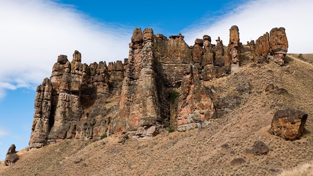 Brown mudflow cliff formations on top of a grassy slope with cloudy blue skies above.