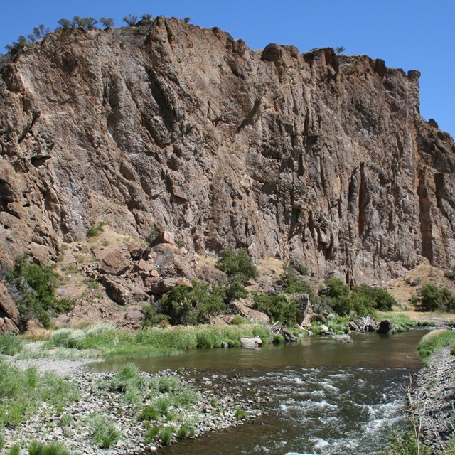 Brown rock formation with green shrubs and a river flowing in front and blue skies above.