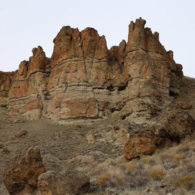 Pillars of brown lahar formations with grassy slopes beneath them and grey skies above.