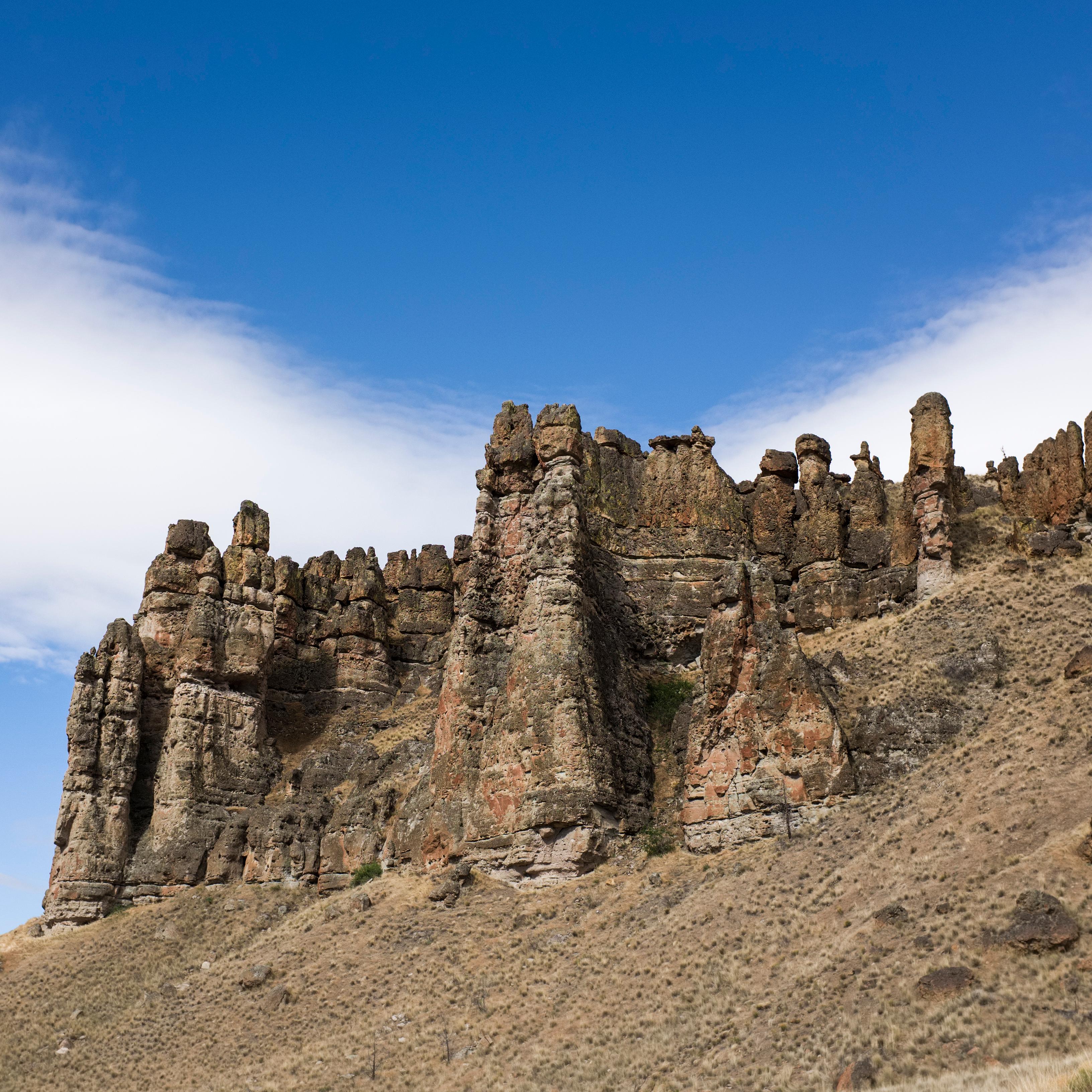 Brown mudflow cliff formations on top of a grassy slope with cloudy blue skies above.
