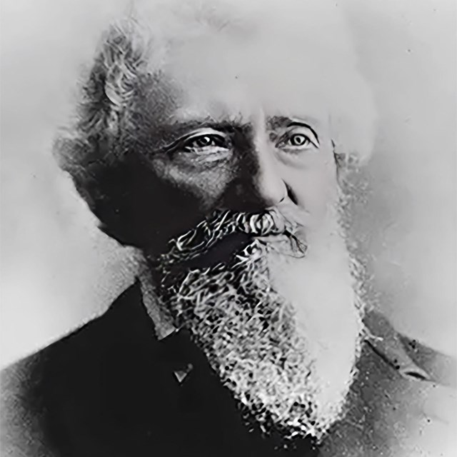 A senior man with a full white beard and hair is dressed in a suit and posing for the camera. 