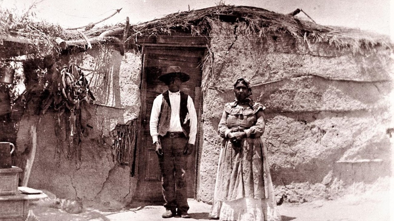 a black and white photo of two people standing in front of a primitive dwelling