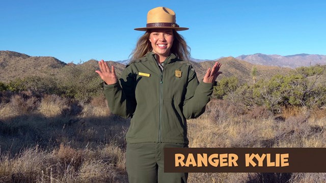 A ranger shrugging her arms, with mountains beyond. Text at the bottom reads Ranger Kylie.