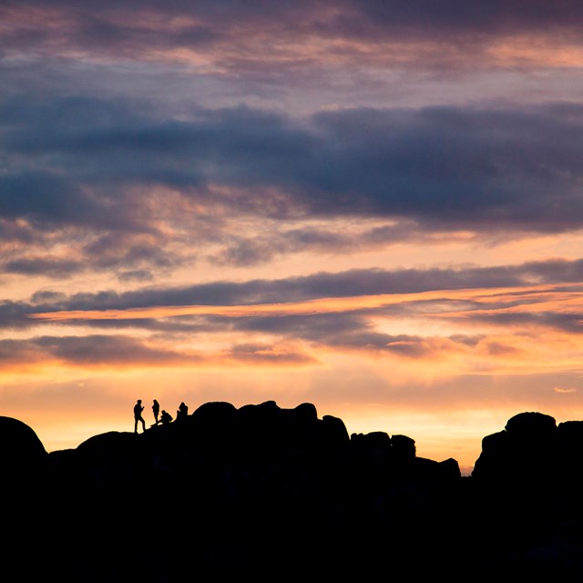 Four individuals atop rock formations silhouetted against a colorful sunset sky.