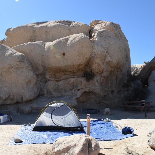 Color photo of a tent site set up in the shadow of a large rock formation. NPS / Hannah Schwalbe