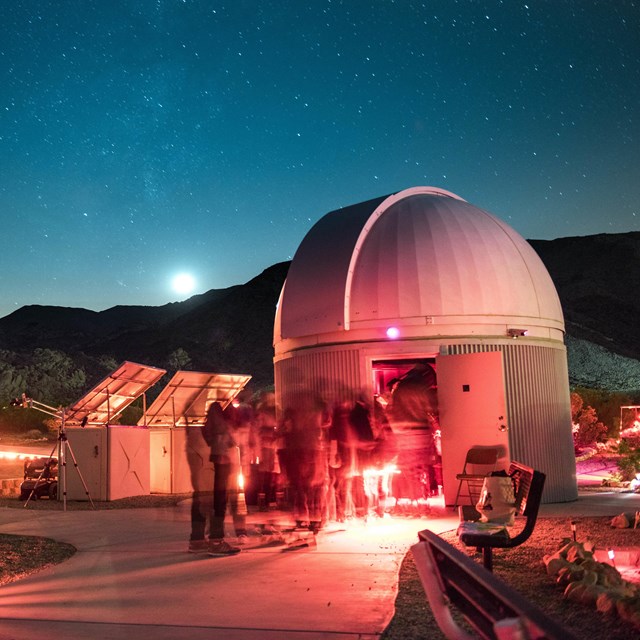 A white observatory building with people around it using red lights and the night sky overhead.