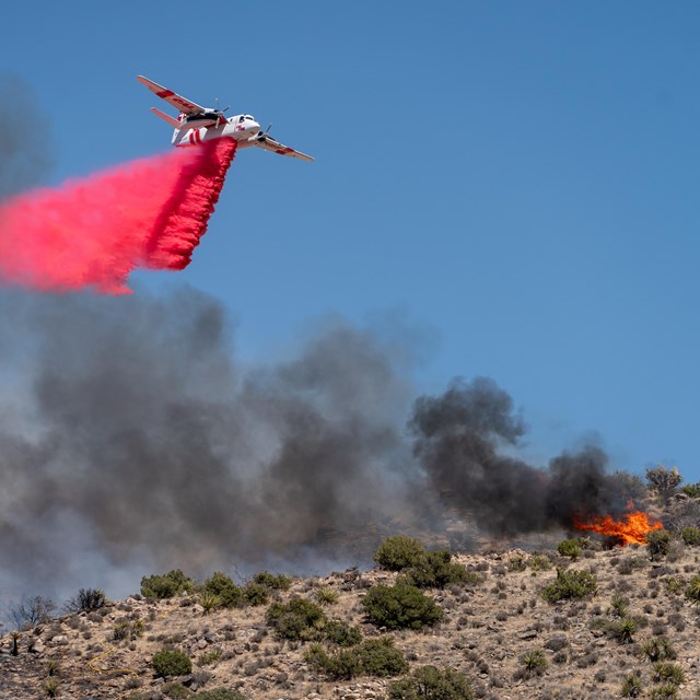 A plane flying over a desert landscape dropping retardant over a fire.
