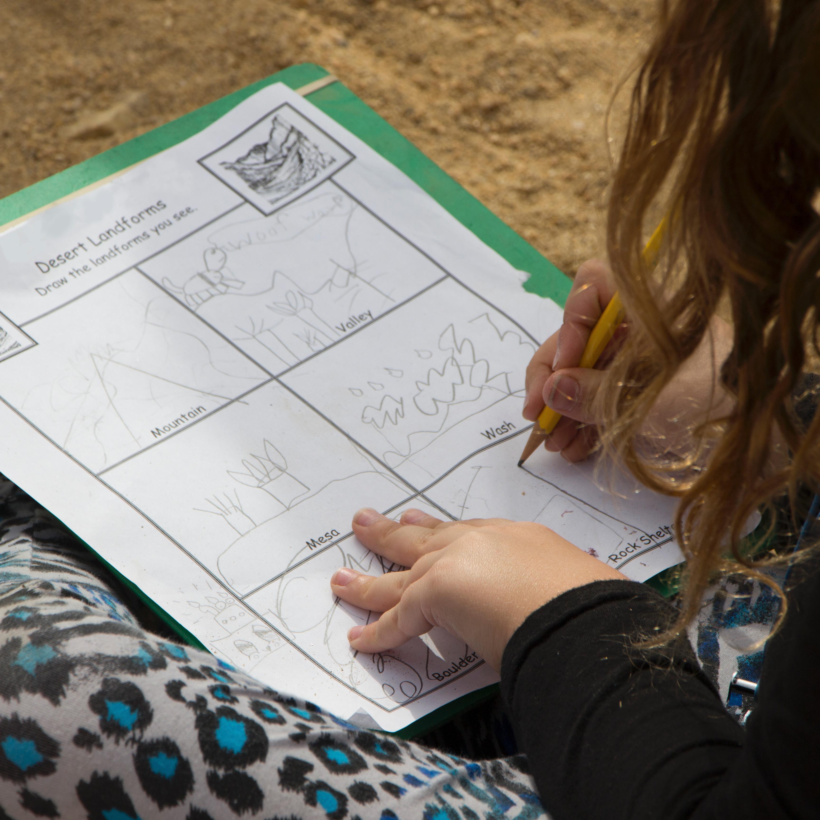 a child sitting on the ground draws desert landforms on a worksheet