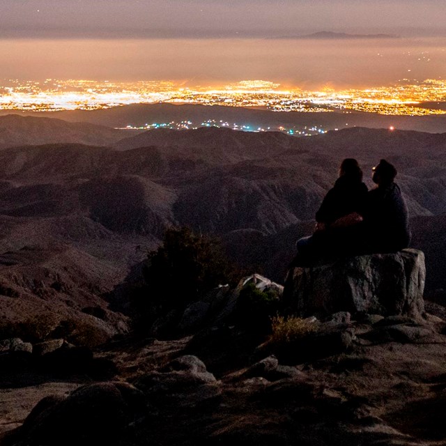 a couple sitting at a viewpoint at night, looking toward the lights of the Coachella Valley