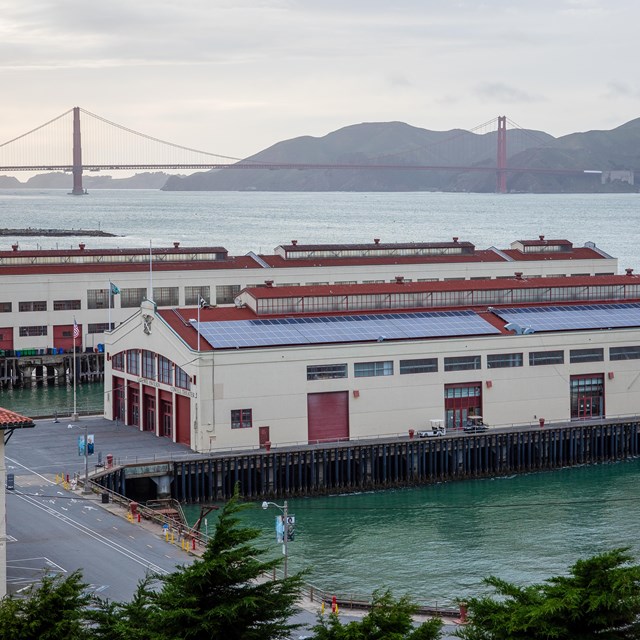 Long, narrow white and red buildings on piers in the San Francisco Bay with the Golden Gate Bridge