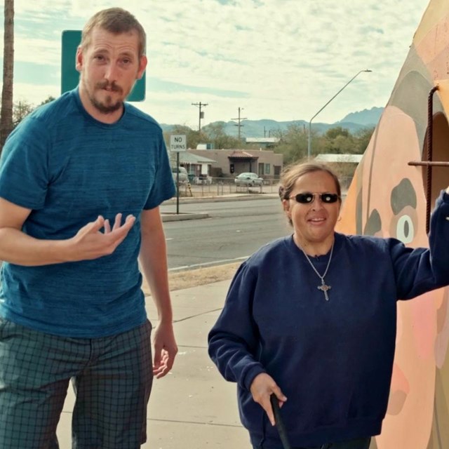 A man signs while a woman with sunglasses and a white cane feels the side of a mural