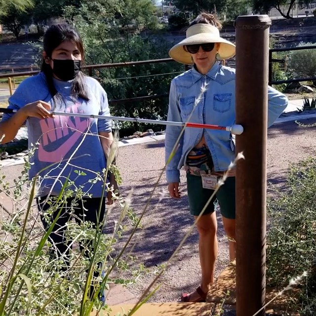 A woman wearing a face mask carrying a white cane touches the cane to a metal structure in a park