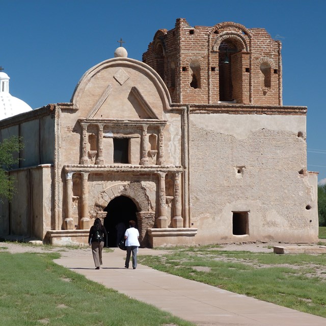 Two people walk into an earth-colored church with a belltower and arched entrance