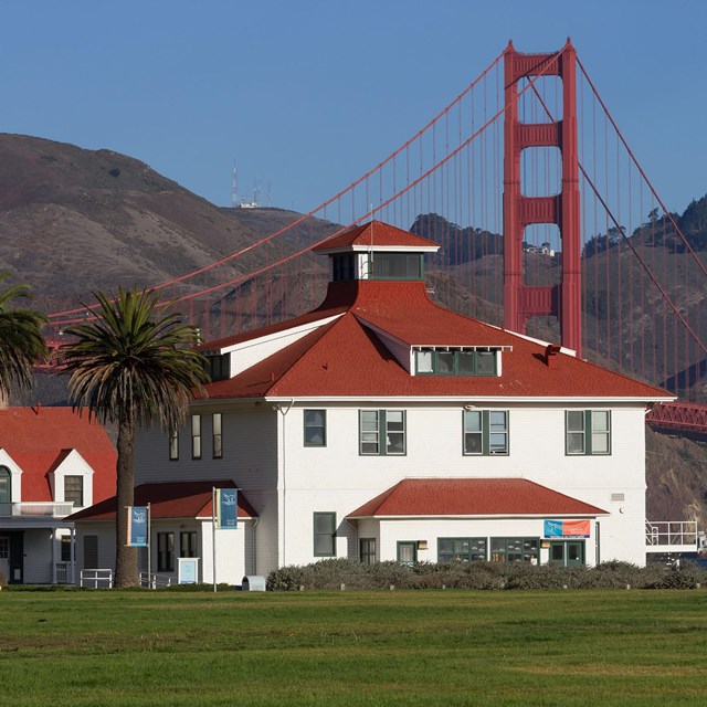 White buildings with red roofs sit on a green lawn with the Golden Gate Bridge in the background