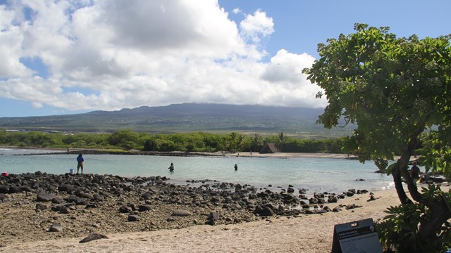 White sand and black lava in foreground, backed by a shallow bay and a distant cloud-covered volcano