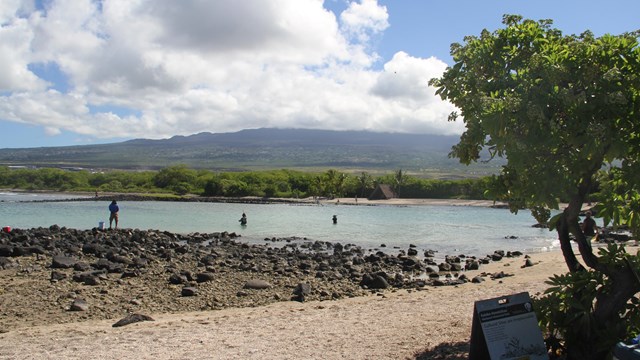 White sand and black lava in foreground, backed by a shallow bay and a distant cloud-covered volcano