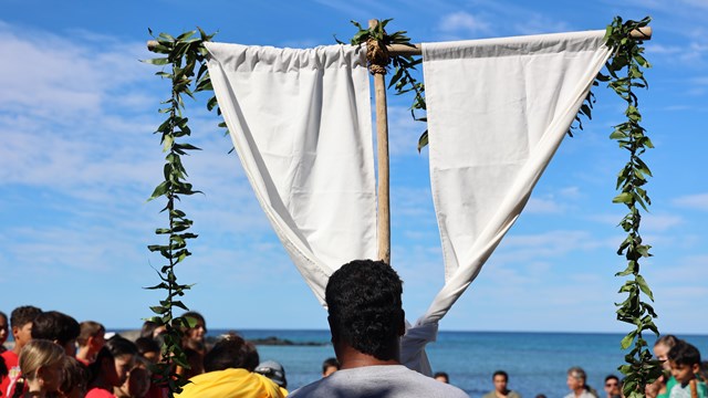 Man carrying traditional Hawaiian pole with lei and white fabric.