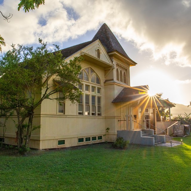 A large tan building, black roof, steeple, and green lawn. The sun is setting behind the building. 