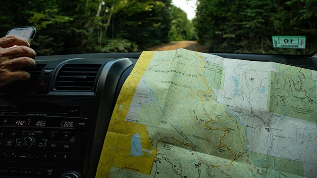  a park map and a hand grasping a phone on the dashboard of a car, trees and a dirt road are visible
