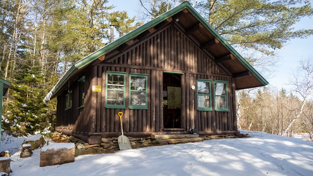 A brown cabin with green trim sits on a snowy piece of land with green trees and a blue sky.