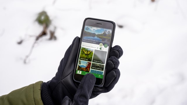 A gloved hand holds a mobile device that shows the NPS app on screen. There is snow in back.