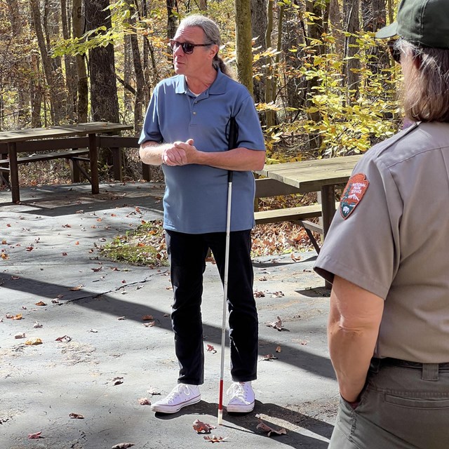 A man wearing sunglasses and holding a white-tipped cane stands with his hands clasped.
