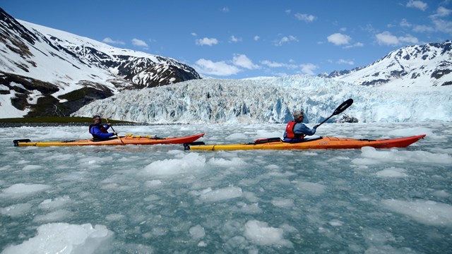 Two kayakers paddle through ice chunks from the background glacier.
