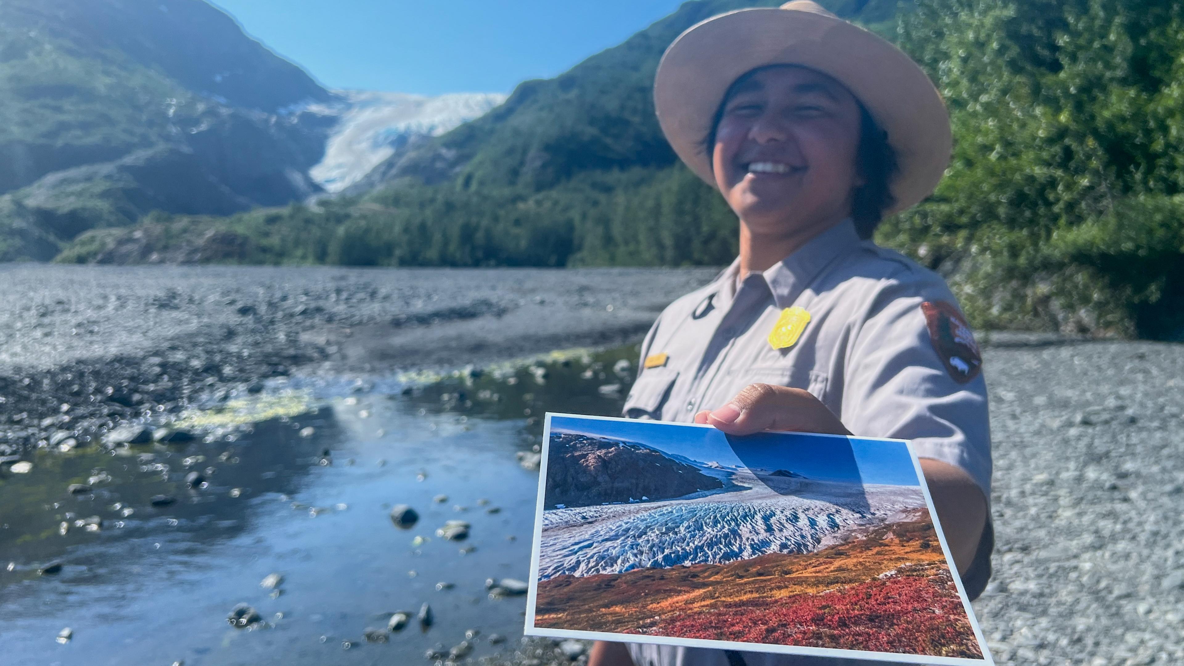 A ranger holds out a postcard with glacier behind