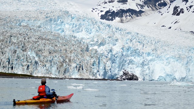 A kayaker on water with a tidewater glacier in the distance.