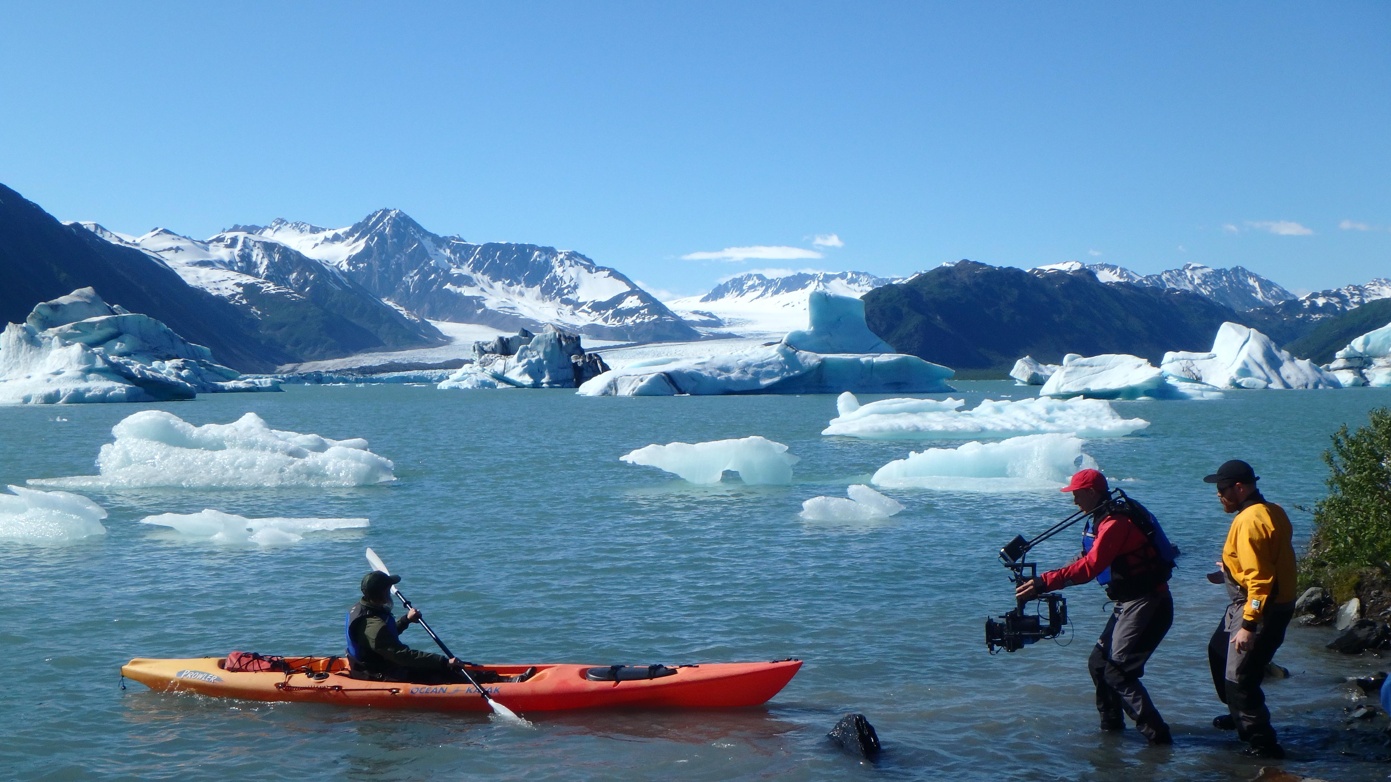 A person films a kayaker on an iceberg filled lagoon.
