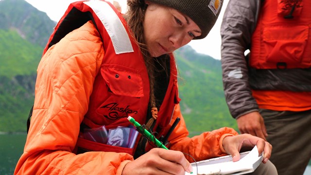 A person in bright orange lifevest writes on paper on a clipboard