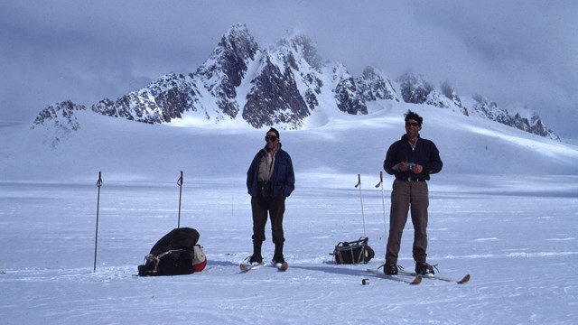 In this historic photo, two people wearing skis and dressed warmly stand on a snow covered icefield.