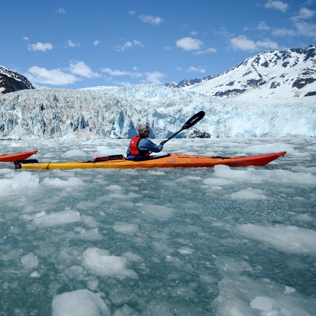 Two kayakers paddle through ice chunks from the background glacier.