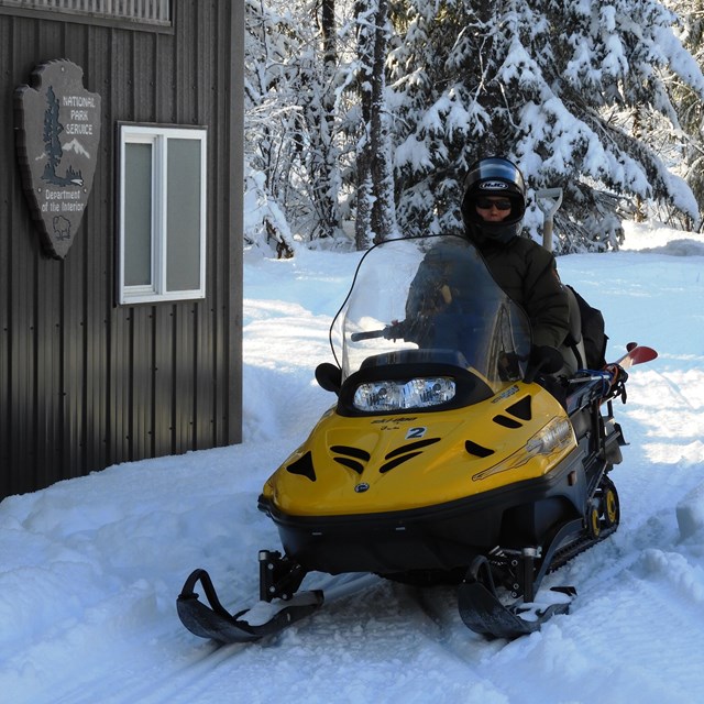 A park ranger sits on a snowmobile in front of a shed. 