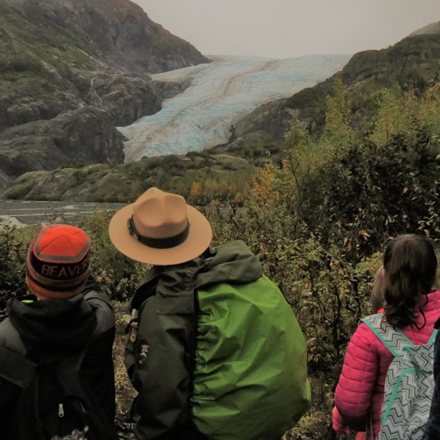 A group of people look at a distant glacier. A Ranger in their flat hat is in middle of the group. 