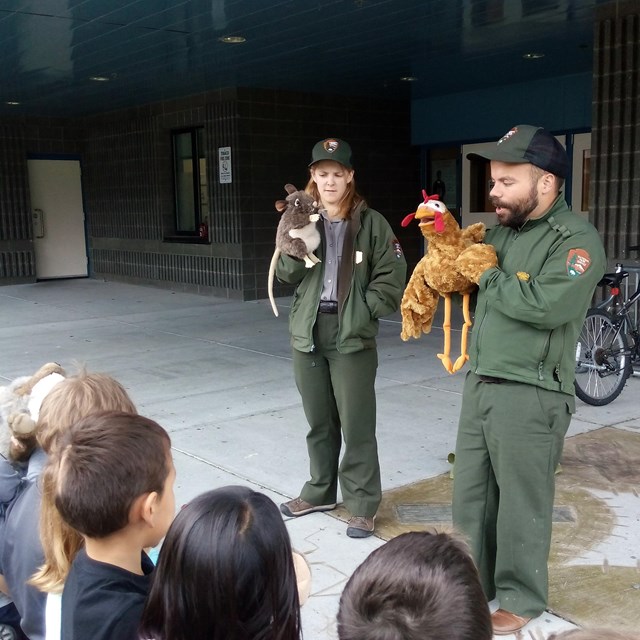 Two Park Rangers talk to a group of students.  The park rangers are holding stuffed animals.