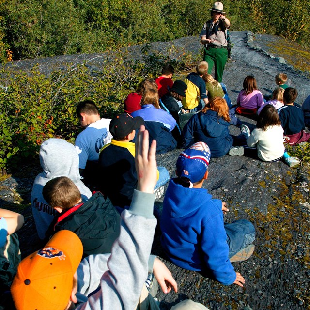 A ranger talking to a group of children. One of the children is raising their hand.