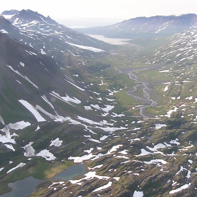 An aerial view of an alpine meadow with pockets of snow, surrounded by mountains.