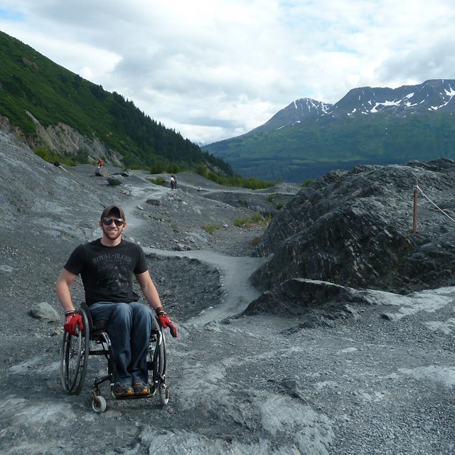 Man in a wheel chair on mountainous rocky area with trail in the background (Edge of Glacier Trail).