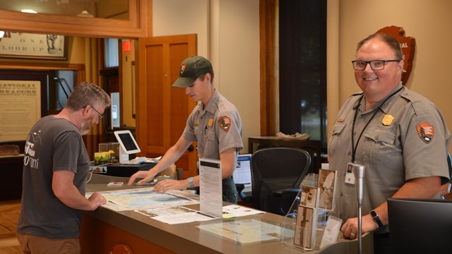 Two park rangers standing behind a desk talking to a park visitor.