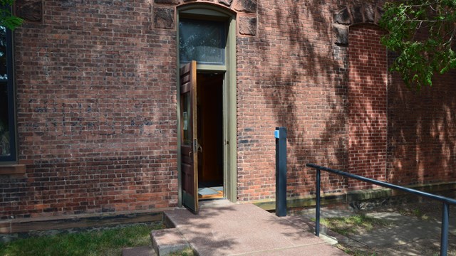 A walkway with a railing on the right leads towards a door to a brick building.