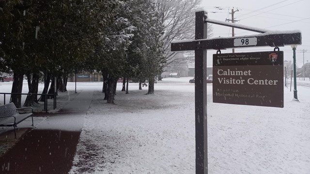 A blanket of white snow covers the ground surrounding a park sign with trees in the background.