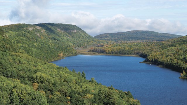 A large lake surrounded by a dense green forest and rolling hills.