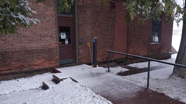 A walkway with a railing on the right leads towards a door to a brick building.