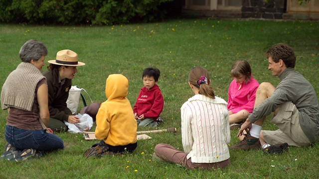 A seated ranger interacts with sitting adults and children on the grass outside.