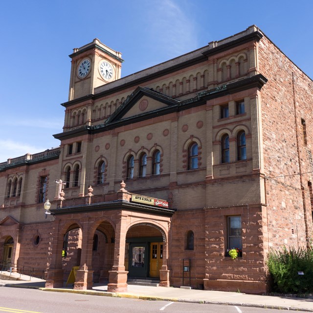 A 3 story red stone building stands along a street, with a tree on the right with blue skies.