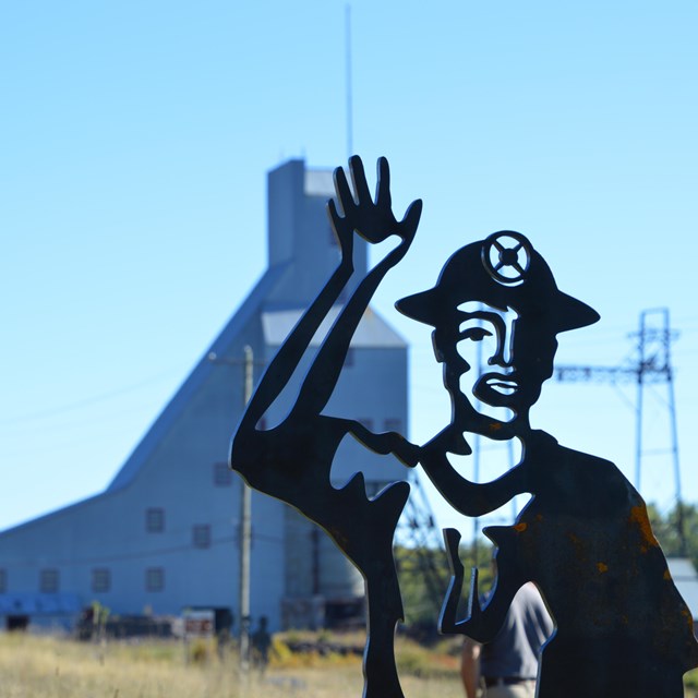 A metal statue of a man waving with a tall mining structure in the background.