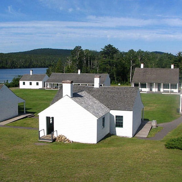 numerous smaller white historic structures on a grassy field. A lake is in the left background.