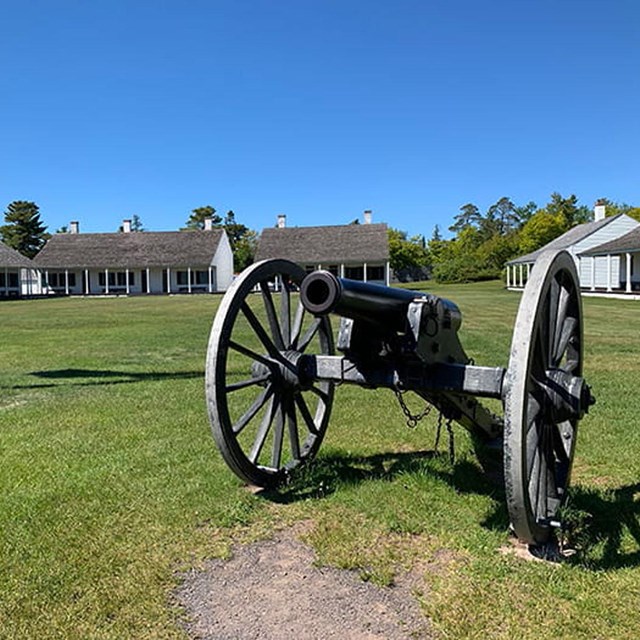 A historic cannon sits on a grassy field, with white wood buildings in the background