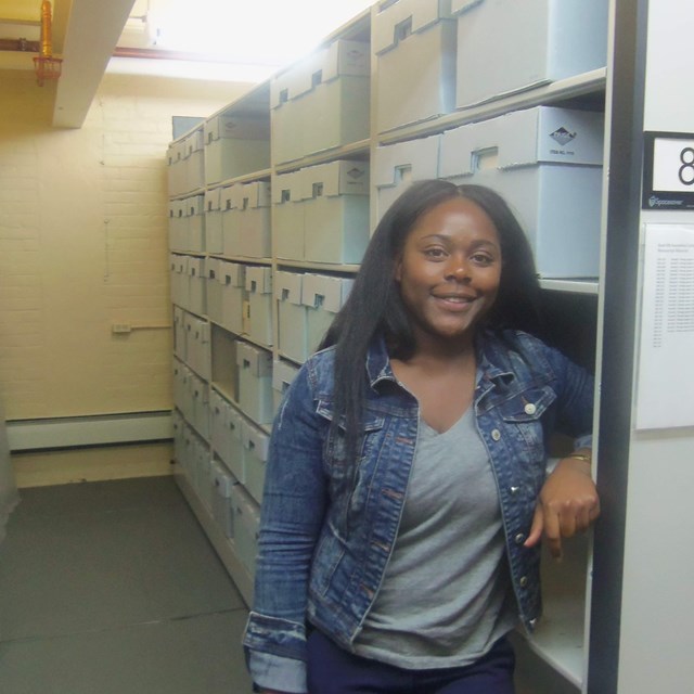 A woman standing next to a wall of shelves full of gray archival boxes.